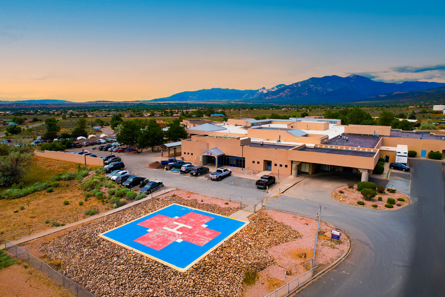 Aerial view of a healthcare facility with a prominent helicopter landing pad and surrounding parking area against a mountain backdrop.