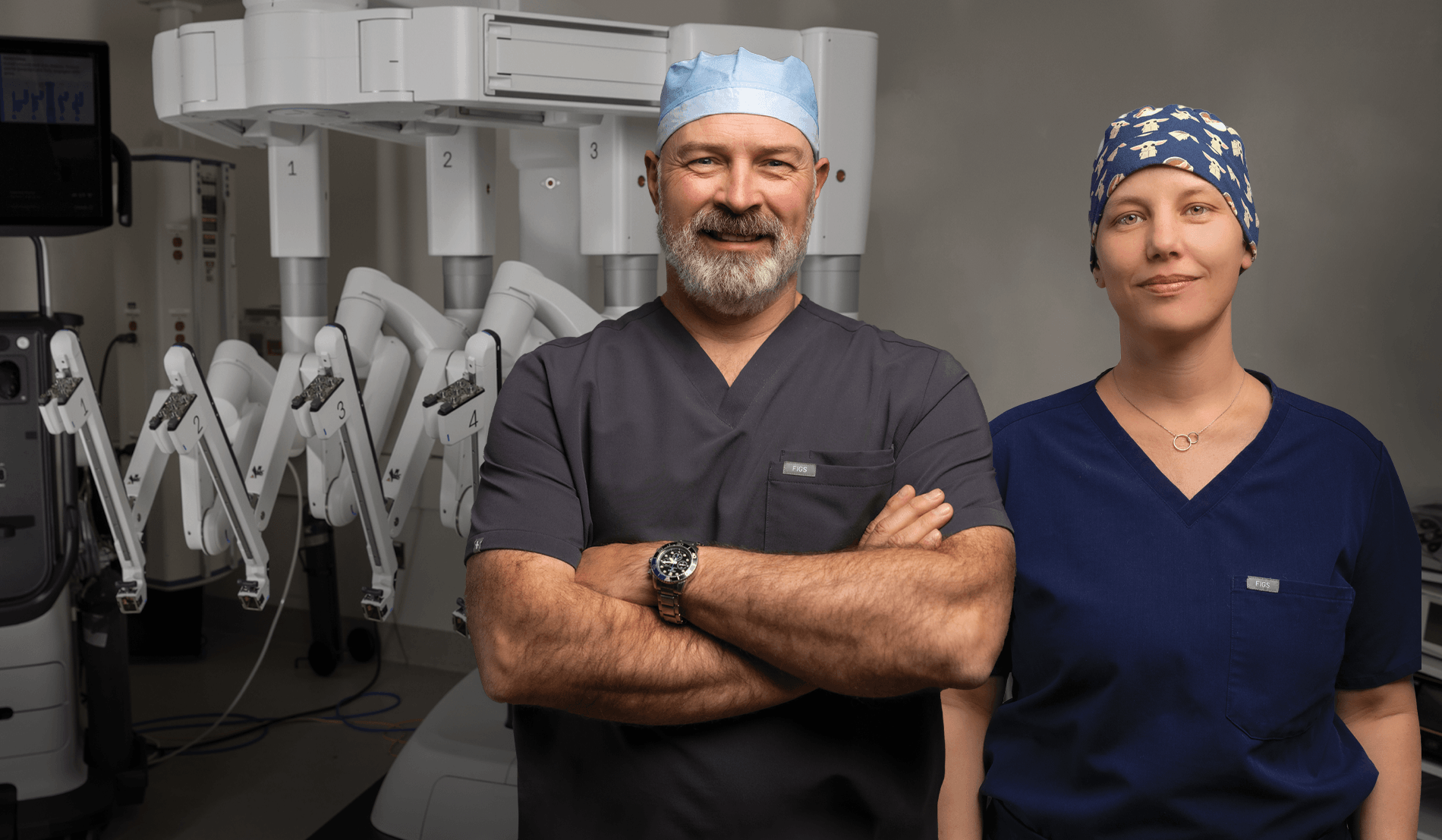 Two medical professionals in scrubs stand confidently in a surgical room with robotic surgery equipment in the background.